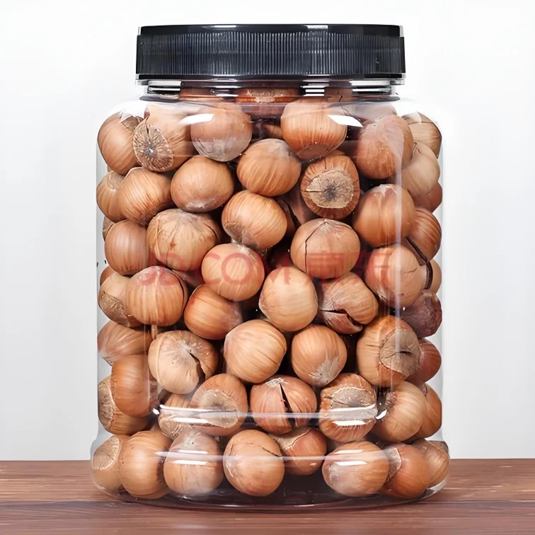A clear plastic jar with a black lid filled with whole hazelnuts in their shells, sitting on a wooden surface against a plain white background.