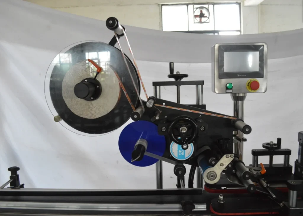 A close-up of a Top and Bottom Labeling Machine with spools and a touchscreen control panel, set against a plain white background in a brightly lit room.