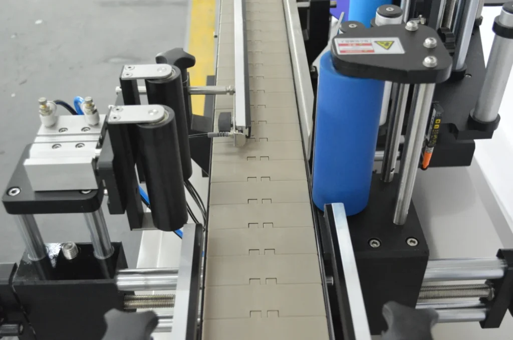 A close-up view of an industrial conveyor belt system with rollers and mechanical arms in a factory setting, featuring blue, black, and metallic components—ideal for integrating equipment like a Round Bottle Labeling Machine.