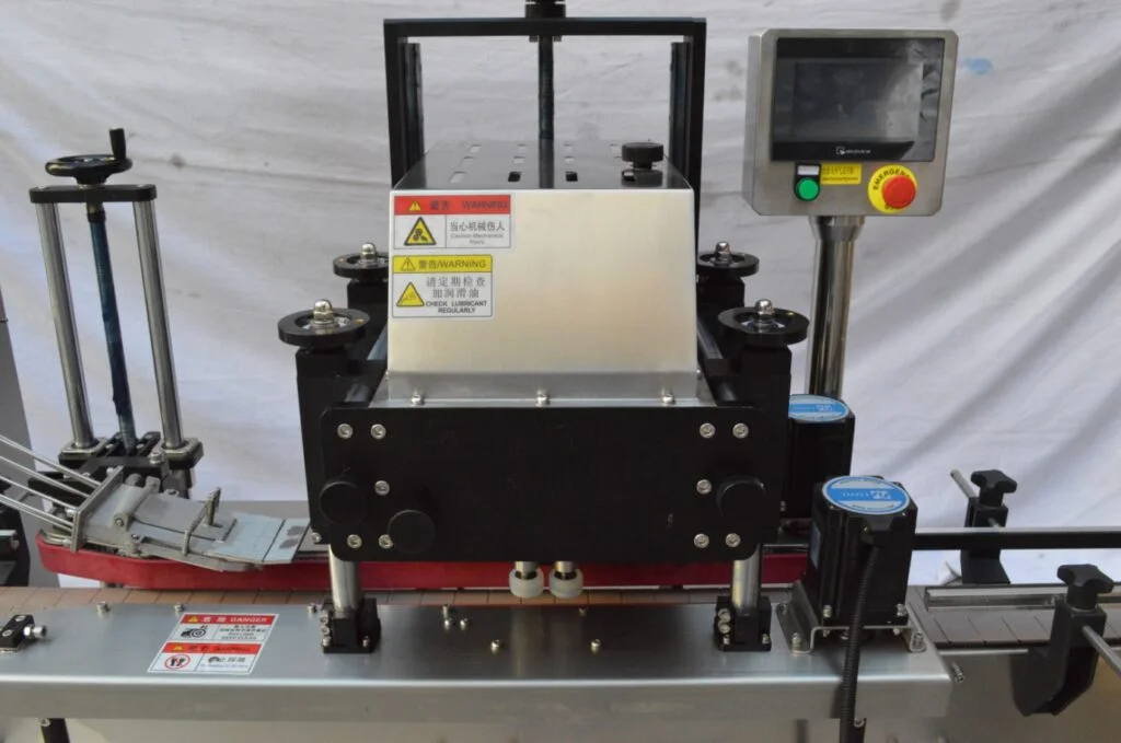 A close-up view of an industrial labeling machine with safety warning labels, a control panel with buttons and a display screen, and a conveyor belt for products.