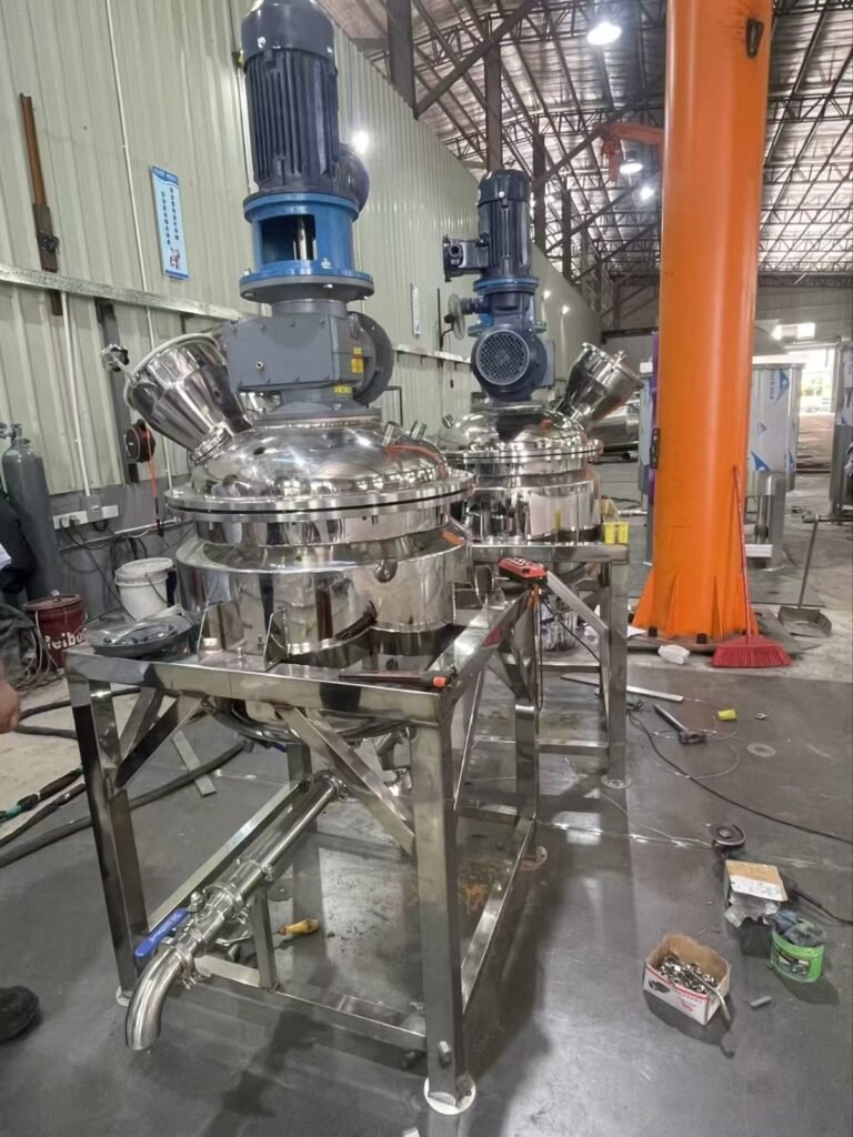 A high-precision stainless steel industrial chemical reactor with a top-mounted vertical agitator motor, integrated pressure gauges, and a bottom discharge pipe, shown inside a manufacturing workshop.