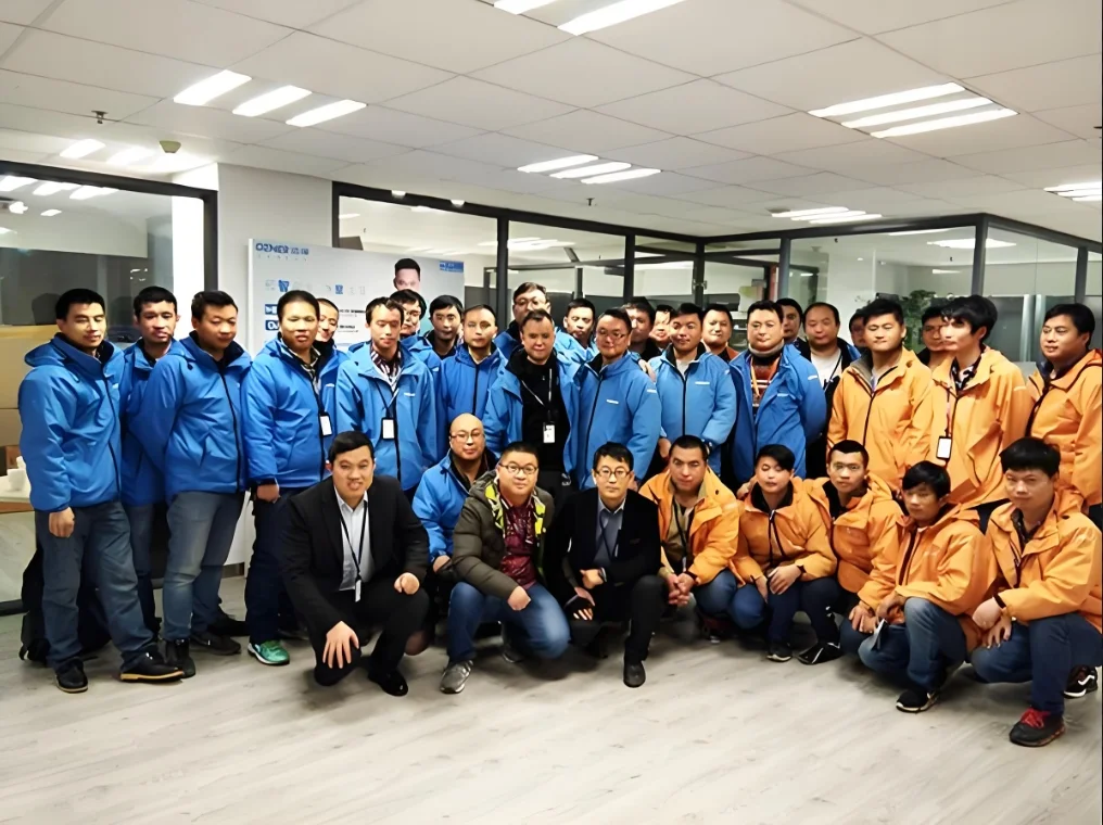 A group of men poses for a photo in an office with glass walls and fluorescent lighting. Most are standing, a few kneeling. Some wear blue or orange jackets with ID badges from the filling machines team, while others wear dark suits.