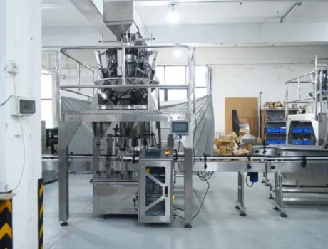 A modern automated packaging machine with a conveyor belt in an industrial setting, surrounded by equipment, boxes, and storage shelves in a bright, clean factory.