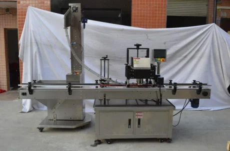 A stainless steel automated labeling machine with a conveyor belt and control panel, set up outdoors in front of a white sheet backdrop and orange tiled wall.