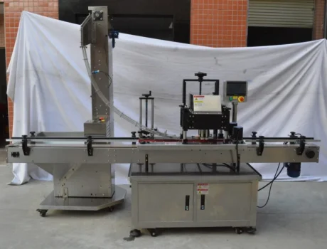 A stainless steel automated labeling machine with a conveyor belt and control panel, set up outdoors in front of a white sheet backdrop and orange tiled wall.